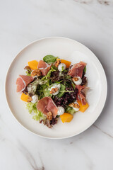 Fresh salad with parma ham, melon slices, walnuts, goat cheese and mixed greens, served on a white plate on a marble table. Restaurant or cafe menu, cookbook recipe

