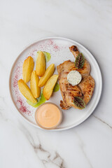 Top view of a delicious grilled chicken served with potato wedges, a creamy sauce, and herbs on a white plate, creating an enticing culinary scene. Restaurant or cafe menu, cookbook recipe

