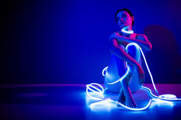 Obraz premium Young woman in neon lights sitting against dark blue background in studio