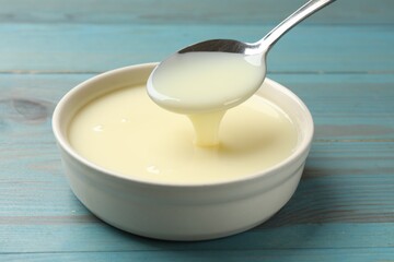 Condensed milk flowing down from spoon into bowl on light blue wooden table, closeup