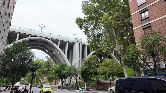 Capture the rhythm of Madrid&rsquo;s streets as vehicles smoothly pass beneath a grand city bridge. This footage highlights the dynamic interplay of urban architecture and daily traffic in the bustling capi