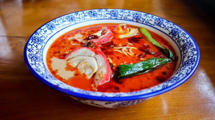 A vibrant bowl of spicy red ramen soup with slices of char siu chicken, chili oil, and green vegetables, served in a blue and white cerramic patterned bowl on a wooden table.
