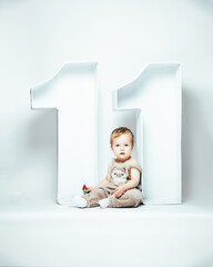 Cute little baby with big numbers eleven on white background. 11 months old child sitting in studio. Blond toddler in overalls and white socks