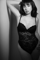 Fototapeta premium Black and white boudoir portrait of a beautiful caucasian woman wearing a lace teddy. She is leaning against a wall. She has dark hair with bangs. 