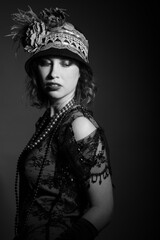 Black and white studio portrait of a caucasian woman dressed in 1920's fashion flapper outfit. She has shoulder length curly hair and is wearing feather hat. 