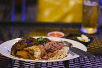 plate of food on a metal mesh table. The plate contains rice topped with what appears to be braised meat and vegetables, garnished with some herbs.