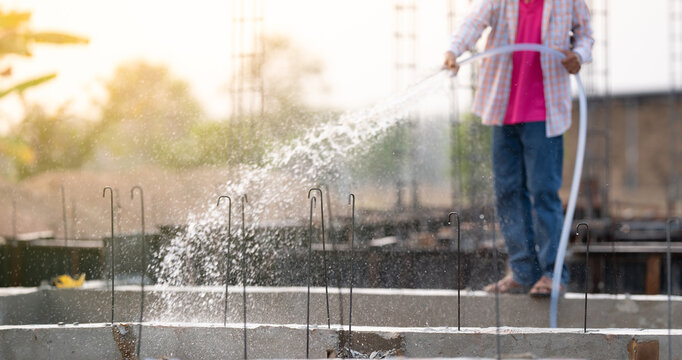 Construction worker watering reinforced concrete beams at a building site, focusing on the curing process for crack prevention, durability enhancement, and protection of reinforced steel structure.