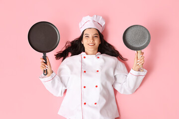 Happy young female cook with frying pans lying on pink background