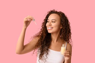 Beautiful young African-American woman with bottle of face serum on pink background