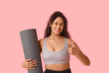 Sporty young African-American woman with yoga mat showing thumb-up gesture on pink background