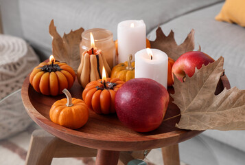 Wooden tray with glowing candles, apples and autumn leaves on glass table in room