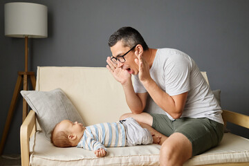 A father playfully engages with his baby on a sofa in a modern living room. The setting conveys warmth and joy as they share a light-hearted moment together