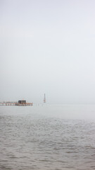 A solitary figure sits on a pier, overlooking a misty expanse of water. Distant structures and a lighthouse emerge from the fog.