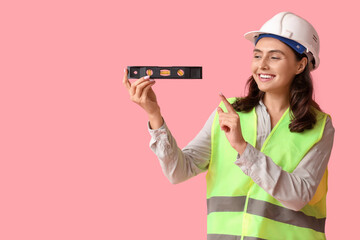 Young female construction worker with bubble level on pink background. Woman in male profession...
