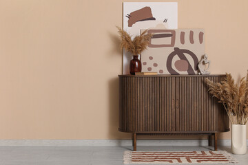 Chest of drawers, pampas grass in vases and pictures near brown wall in room
