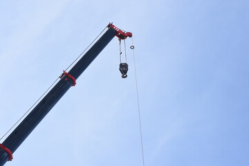the upper part of a crane against a clear blue sky. The crane's arm is extended diagonally, and a hook is hanging from the end of the crane's cable.