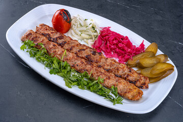 Two skewers of juicy kebabs served with grilled tomato, fresh parsley, marinated onions, pickles, and red cabbage on a white plate. Authentic Middle Eastern food presentation