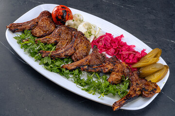 Deliciously grilled lamb chops served with a variety of sides on a white plate. Garnished with fresh parsley, accompanied by grilled tomato, onions with herbs, pickled red cabbage, and pickles.