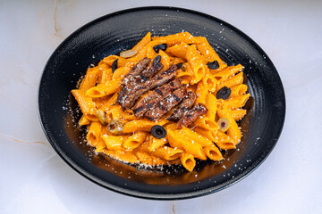 Black plate of penne pasta in creamy orange sauce, topped with grilled beef slices, black and green olives, and grated cheese. Vibrant, appealing, perfect for showcasing a delicious pasta recipe.