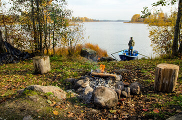 The shore of a lake in autumn. In the foreground there is a fire burning, there are seats made of wooden logs. In the background there is a figure of a fisherman standing in a boat with a fishing rod.