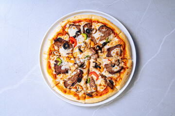 Whole pizza with beef slices, black olives, green and red bell peppers, mushrooms, and melted cheese. Placed on a white plate with a light gray patterned background.