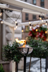 
Close-up of an outdoor oil lamp with flame, string lights above, blurred plants, and a cozy urban background, evoking warmth