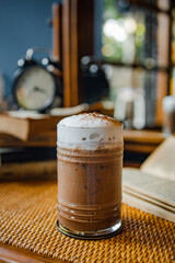 A frothy chocolate drink in a glass, set on a textured surface, with a cozy background featuring a clock and books.