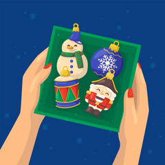 Vector illustration of a hand holding a box with Christmas ornaments snowman, nutcracker, drum, ball.