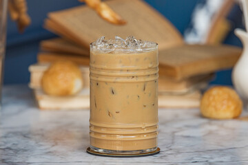 A refreshing glass of iced coffee sits on a marble surface, surrounded by baked goods and open books, creating a cozy ambiance.