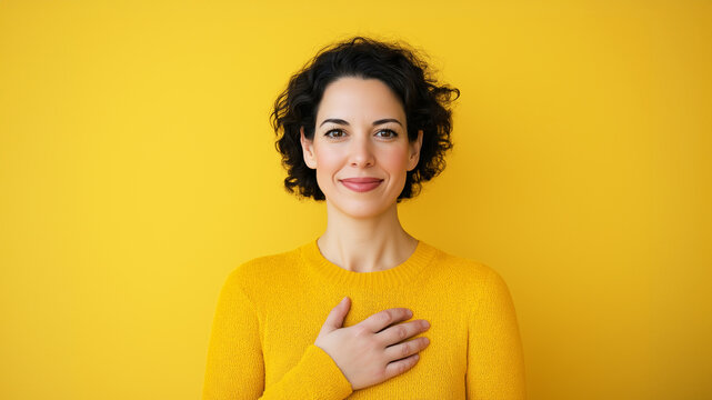 Woman touching her heart area and looking with empathy. Yellow background.