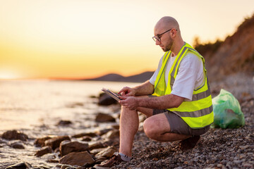 Inspection environmentalist. Side view of adult Caucasian man wearing glasses using tablet. In background is ocean and sunset. Concept of environmental protection
