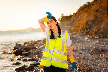 Tired young woman volunteer wearing vest and gloves wiping sweat from forehead. Clean up day....