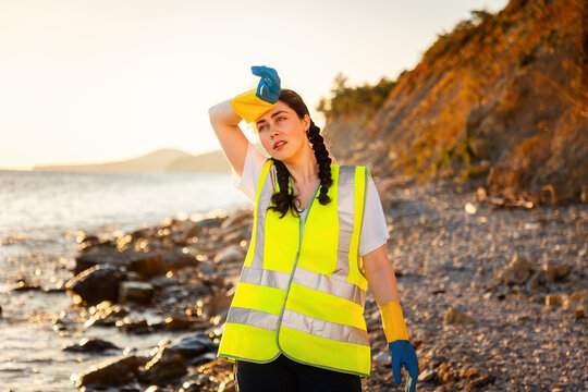 Tired young woman volunteer wearing vest and gloves wiping sweat from forehead. Clean up day. Concept of ocean's pollution and environmental conservation