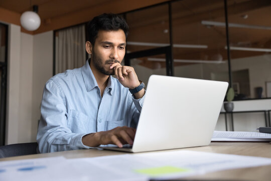 Indian businessman staring at laptop screen deeply focused on his work seated in workplace, analyze received document, review data, consider decision, working on financial report and business strategy - Powered by Adobe