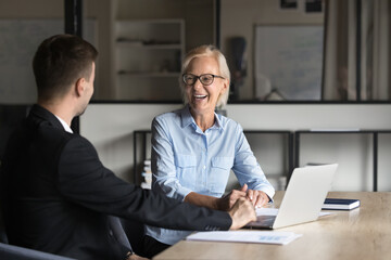 Two colleagues, aged woman and young man engaged in positive professional conversation met in company office with laptop, laugh, discuss positive outcome, project completion, business-related topics