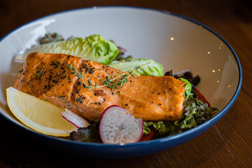 A beautifully plated salmon fillet with herbs, served on a bed of fresh greens and garnished with lemon and radish slices.