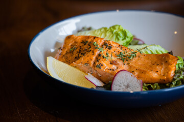 A beautifully plated salmon fillet garnished with herbs, served on a bed of greens with lemon and radish slices.