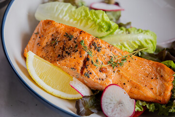 A beautifully plated salmon fillet garnished with herbs, served on a bed of fresh greens and lemon wedges.