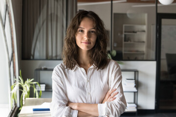 Head shot portrait of pretty young businesswoman posing with arms crossed in modern workspace,...