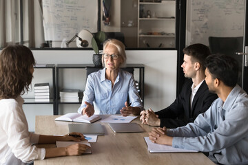 Confident mature 55s female business leader talk to diverse team, take part together in meeting,...