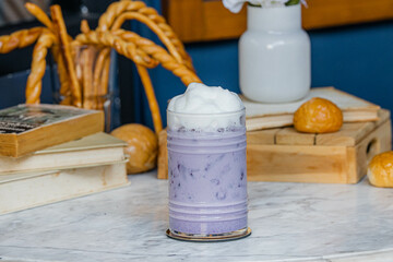 A refreshing purple drink topped with foam, set on a marble table surrounded by baked goods and a vibrant background.