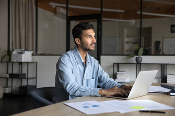 Pensive businessman, entrepreneur working on-line using laptop seated at workplace desk in modern...