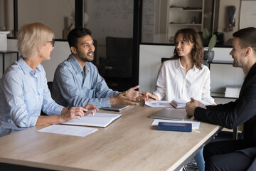 Group of four professionals engaged in strategic discussion, brainstorm, share ideas, listen speech of Indian colleague during meeting around conference table. Collaboration, decision-making, teamwork