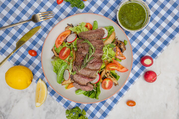 A colorful plate of grilled steak served on a bed of fresh salad, accompanied by tomatoes, radishes, and a vibrant green sauce on a checkered tablecloth.