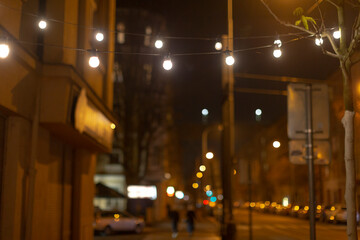 A slightly blurred picture captures a street at night, showcasing a string of warm lights beautifully hanging from a tall pole