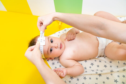 Baby health check with measuring tape indoors. A caregiver measures the head circumference of an infant lying on a colorful mat in a bright, cheerful room.