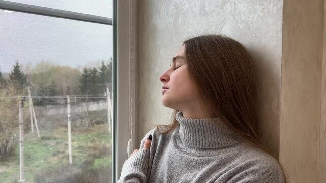 Young woman embracing herself while gazing out the window on a cloudy day, reflecting feelings of sadness and solitude