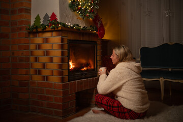 A woman with a mug in her hand sits in front of a fireplace with Christmas decorations