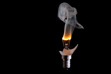 gebrochene Glühlampe mit durchbrennender  Glühwendel, und rauchendem Wolframdraht vor schwarzen Hintergrund und komplett sichtbaren Lampengewinde E29