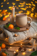 Christmas hot chocolate with tangerines and burning candles on a stack of knitted sweaters creates a cozy winter atmosphere with warm bokeh lights